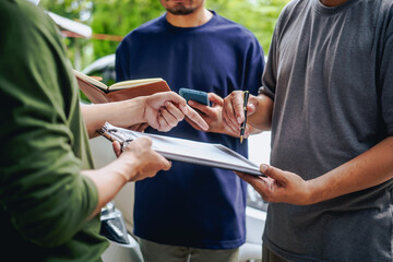 Asian clients engage with a car insurance agent beside a wrecked vehicle. They examine the policy details, assess the damage, and finalize paperwork related to the insurance claim and repairs.