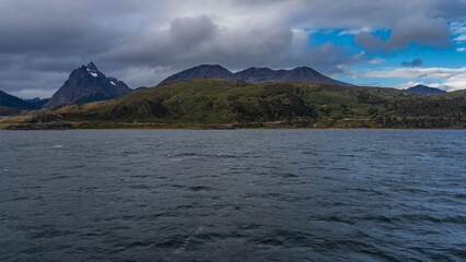 A beautiful mountain range is visible from the ocean. A snow-covered peak against a blue sky and clouds. Ripples on the surface of the water. The Beagle Channel. The Andes. Argentina. Patagonia