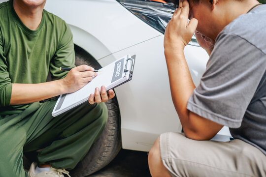 Asian clients engage with a car insurance agent beside a wrecked vehicle. They examine the policy details, assess the damage, and finalize paperwork related to the insurance claim and repairs.
