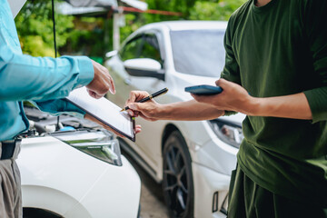 Asian clients engage with a car insurance agent beside a wrecked vehicle. They examine the policy...