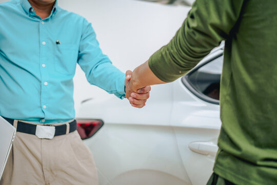 Asian clients engage with a car insurance agent beside a wrecked vehicle. They examine the policy details, assess the damage, and finalize paperwork related to the insurance claim and repairs.