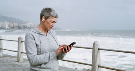 Mature woman, phone and texting on promenade with earphones, reading and workout in morning....