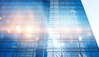 Obraz premium A low-angle view of a modern skyscraper with a glass facade, reflecting the sun and the surrounding urban environment.