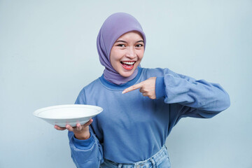 A smiling Asian Muslim woman is fasting and hungry and holding and pointing to a plate.