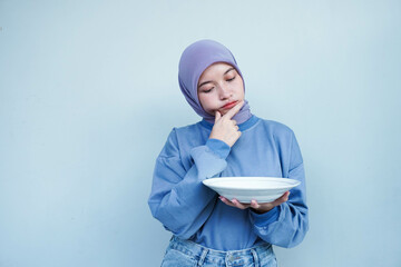 Asian Muslim woman is fasting and hungry and holding cutlery while looking a plate thinking about what to eat with empty plate.