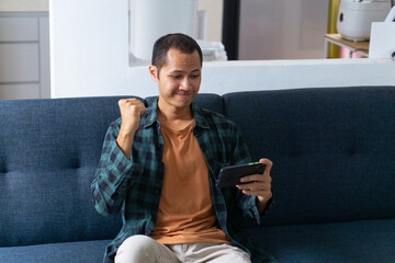 Young asian men wearing orange shirt and flannel make fist gesture while playing mobile game on sofa