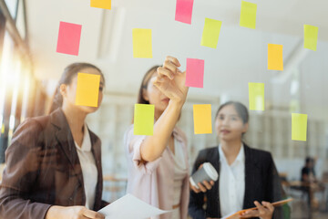 Asian businesswoman teamwork planning with sticky notes and new ideas on a glass wall, Business people brainstorming strategy and new ideas concept, startup worker meeting,