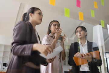 Asian businesswoman teamwork planning with sticky notes and new ideas on a glass wall, Business people brainstorming strategy and new ideas concept, startup worker meeting,