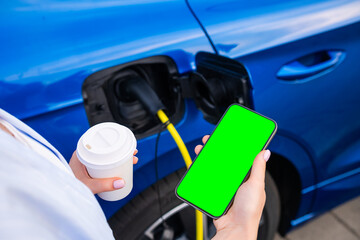 Electric car is being charged at a station while the driver holds coffee and a smartphone