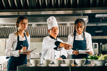 Asian senior chef in uniform teaches cooking techniques to multiracial students. Emphasizing teamwork learning and note-taking in this professional kitchen workshop. Food Edocation