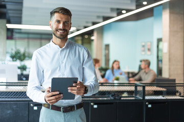 Smiling entrepreneur is standing in office with digital tablet and looking at camera. On the background people doing their work.