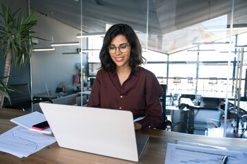 Smiling young middle eastern business woman specialist working focused on laptop computer. Middle aged latin hispanic investor, bank worker using pc for analysing, network at workplace desk in office