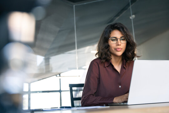 Focused young professional it specialist latin hispanic business working on laptop pc sitting at desk in modern office. Middle eastern indian woman using computer technology app for work. Copy space