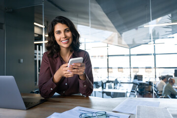 Young Indian business woman manager ceo using cell phone mobile app. Portrait at camera Latin...