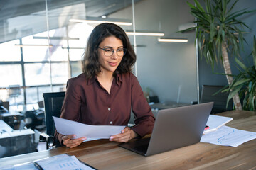 Focused latin hispanic young business woman working on laptop computer reading financial document...