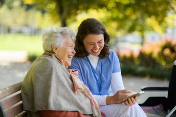Portrait of nurse and elderly woman on walk in the park during warm autumn day. Young caregiver...