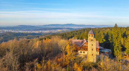 A stunning autumn morning shows the Bramberk lookout tower and nearby hut nestled within the vibrant Jizera Mountains, surrounded by colorful fall foliage and clear blue skies.