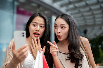 Two excited Asian female friends looking at a smartphone together, amazed by the big sale.