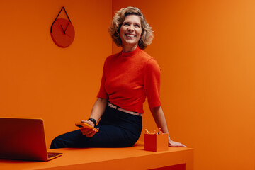 Happy mature woman smiling in a vibrant monochromatic orange office setting, conceptual studio portrait