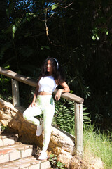 Young, beautiful, Latin, brunette woman with curly hair, dressed in sports clothes, listens to music with white headphones while leaning on the wooden fence after a hard workout.