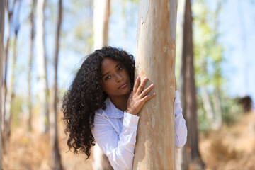 A young, beautiful, Latin, brunette woman with curly hair is holding on and leaning out from behind the trunk of a eucalyptus tree. The woman makes different expressions. Tree hugging concept.