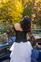 Girl in black leather jacket, white long puffy skirt and leather boots stands next to blue car in...