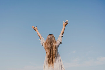 A woman with long hair is standing on the beach, looking up at the sky. She is wearing a dress and she is happy.