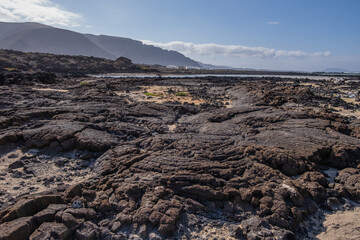 White sand beach with solidified volcanic lava flows, black rocks. Mountains in the background and sky with big white clouds. Caletón Blanco, Lanzarote, Canary Islands, Spain