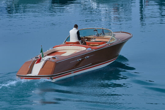 Luxury varnished wooden boat with white leather chairs, carrying a man across vibrant blue water. Exclusive, high-end boat in motion, showcasing polished wood and elegance.