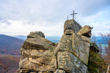 Fototapeta premium The rocky Oresnik formation features a lookout point and wooden summit cross, offering panoramic views of the Jizera Mountains in Czechia on a cloudy day.