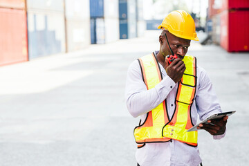An African construction worker in a safety vest and hard hat checks his tablet while talking on walkie talkie, surrounded by containers in an industrial area. Shipping business management.