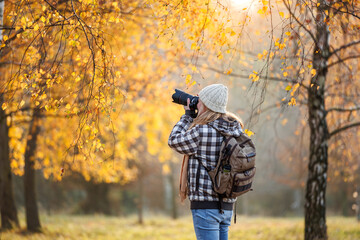 Woman hipster with camera taking picture of autumn leaf. Tourist hiking in birch forest. Landscape photographer with backpack enjoying nature in fall season