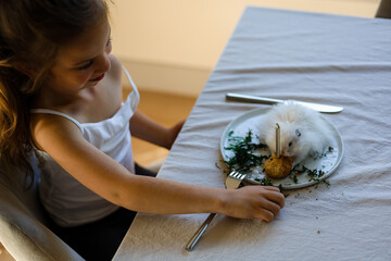 Little girl sitting at the table and look tho the dinner plates with hamster inside that eating pastry and grasses.
