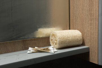 Bathroom shelf with a rolled-up loofah sponge and wooden cotton swabs. The background features a wooden surface and a foggy mirror, emphasizing an eco-friendly and natural hygiene setup