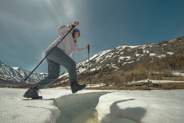 A plus-size girl with trekking poles jumps over the pripyat. plus-size traveler is a tourist. Jump over the winter river. A river in the snow against the background of mountains and a tourist.