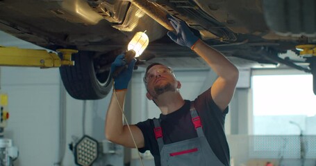 Car service - mechanic check out automobile parts while working with lamp. Auto mechanic working underneath car lifting machine at the garage.