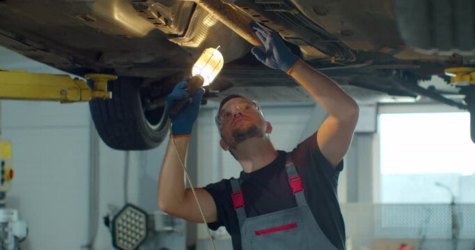Car service - mechanic check out automobile parts while working with lamp. Auto mechanic working underneath car lifting machine at the garage.