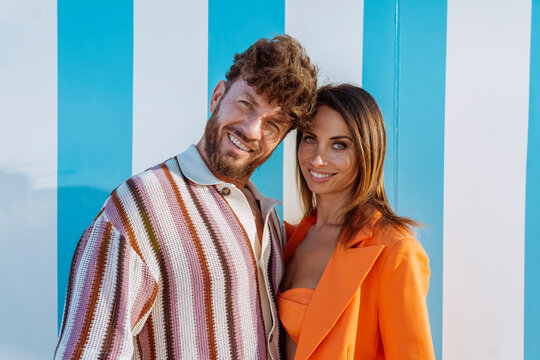 Fashionable couple posing together happily in front of a blue and white striped wall.