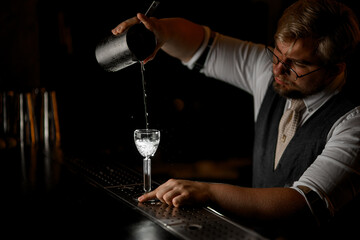 Male bartender in glasses pours a clear drink from a mixing glass into a stemmed glass