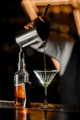 Selective focus of stemmed glass into which bartender pours cocktail from metal mixing glass