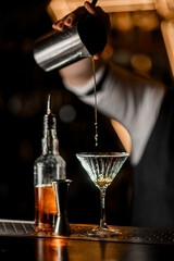 Selective focus on stemmed glass into which male bartender hand pours cocktail from metal mixing glass