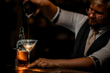 Side view of smiling male bartender pouring cocktail prepared in mixing glass into stemmed glass
