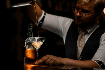 Male bartender pours a cocktail prepared in a mixing glass into a glass on a tall stem
