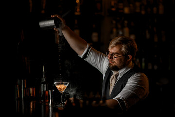 Smiling bartender pours the last drops of the finished drink into a glass on a tall stem