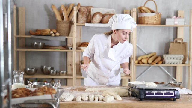 Skilled young female baker in white chef coat and hat carefully slicing and weighing pieces of dough on flour-covered worktable in cozy bakery filled with freshly baked bread and baking tools