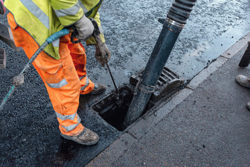 Construction worker operates a vacuum truck to clear debris from a storm drain on a busy city...