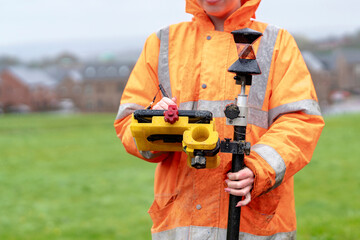 Surveyor in bright orange rain gear measures land using surveying tools on a cloudy day in an open...