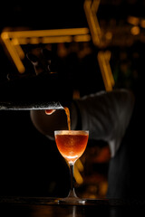 Bartender's hand finishes filling a frosted stemmed glass with an orange cocktail by pouring it...