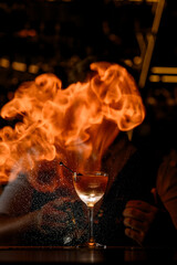 Against the background of a stemmed glass filled with a golden cocktail bartender sprays a flammable liquid in the air and ignites it