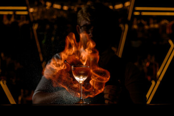 Glass with a transparent drink is illuminated by a flame, which is lit by a bartender in its background
